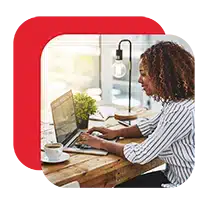 Woman working on a laptop at a wooden desk with coffee and a small plant.